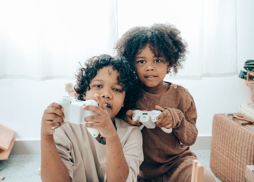 Free Adorable African American sisters in domestic clothes sitting on floor at home and playing video games with gamepad while spending time together and looking at camera Stock Photo