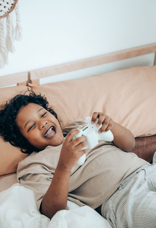 Free From above of laughing cute African American child in pajama lying on cozy bed and having fun while playing video games during spare time at home Stock Photo