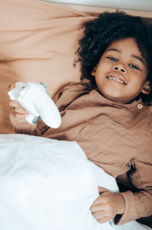 Free From above of smiling little African American child in sleepwear lying in cozy bed and using gamepad while spending free time at home and looking at camera Stock Photo