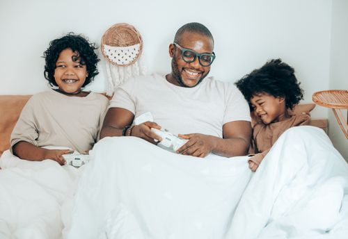 Free Smiling adult African American man wearing glasses lying in comfortable bed with cute daughters in pajamas and using gamepads during free time at home Stock Photo