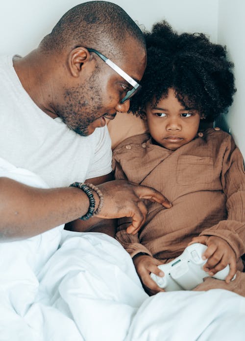 Free Tender adult African American man wearing eyeglasses talking with adorable girl while lying in bed and using joystick for video games in weekend Stock Photo