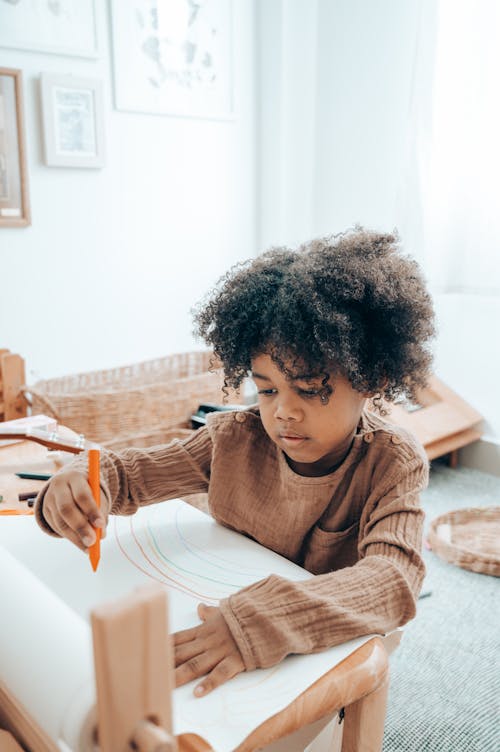 Cute focused African American girl in domestic clothes sitting at wooden table and drawing with marker on paper sheet while spending free time in modern apartment