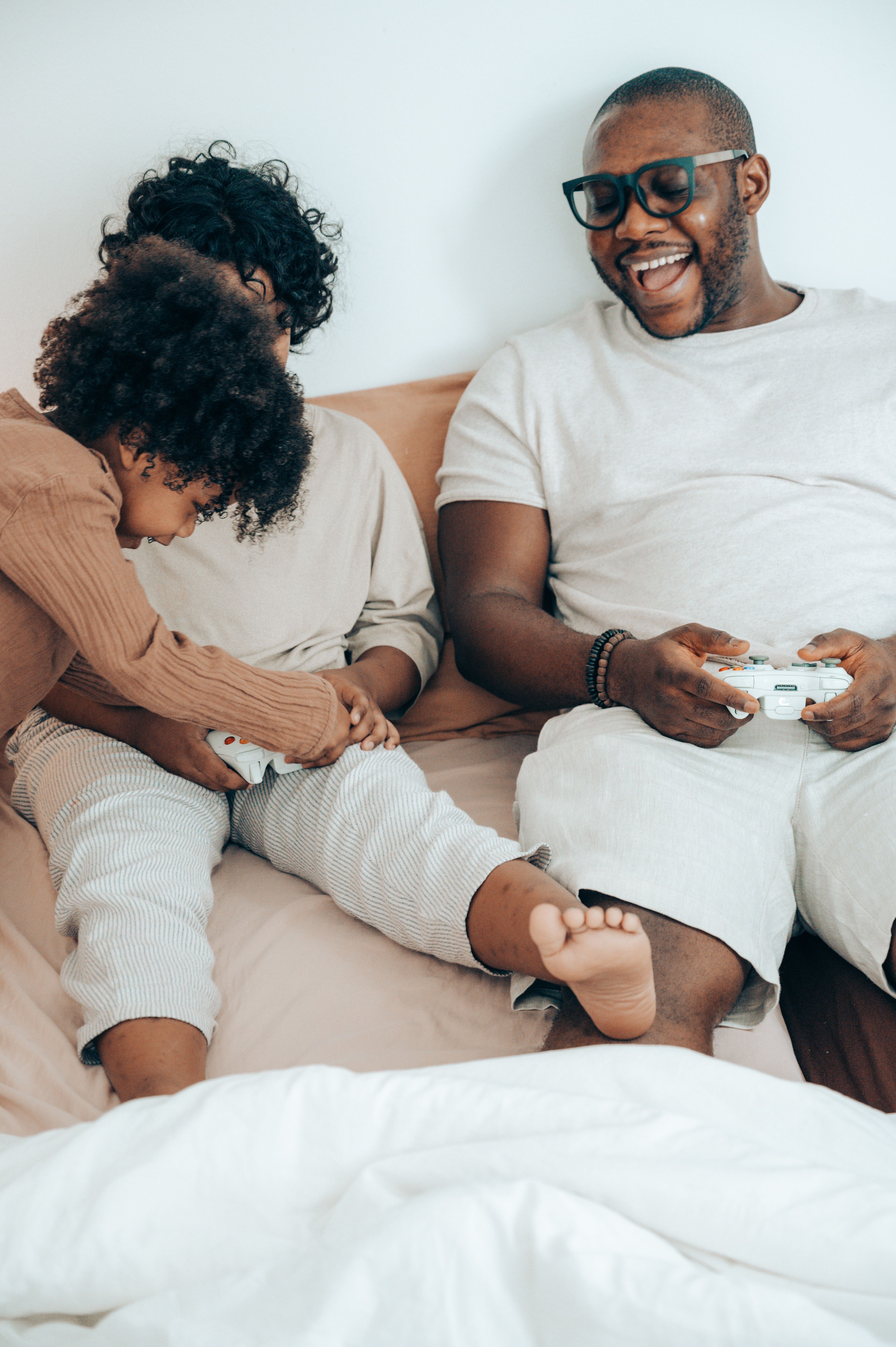 Laughing bearded African American man in eyeglasses lying on bed and using joystick while spending free time together with adorable kids