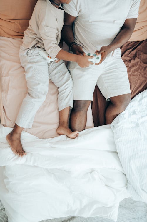 Top view of unrecognizable African American man and kid lying together on comfortable bed and using gamepad while resting in modern bedroom