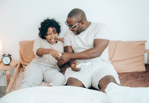 Black father and kid using joysticks on bed