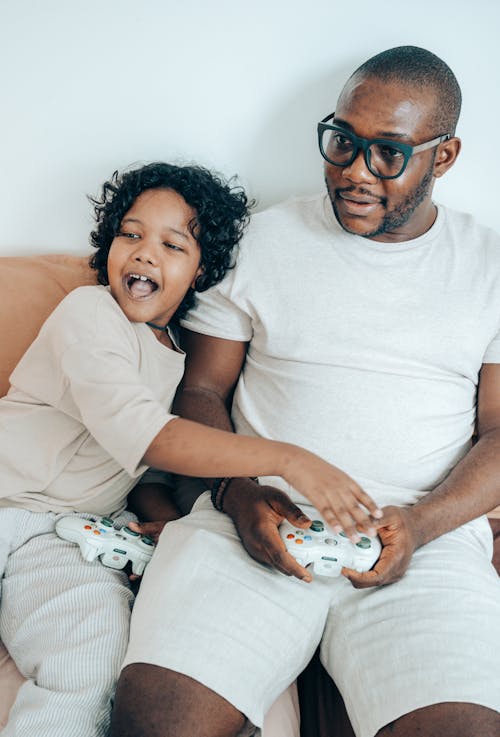 From above of cheerful young man in glasses with kid enjoying play on game console while relaxing together on sofa in cozy apartment