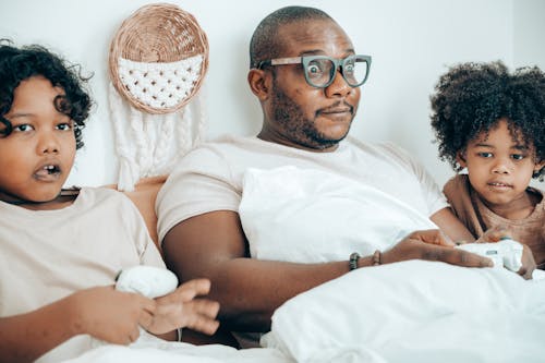 Cheerful young black man in glasses sitting with kids on bed in comfortable bedroom and playing together during weekend