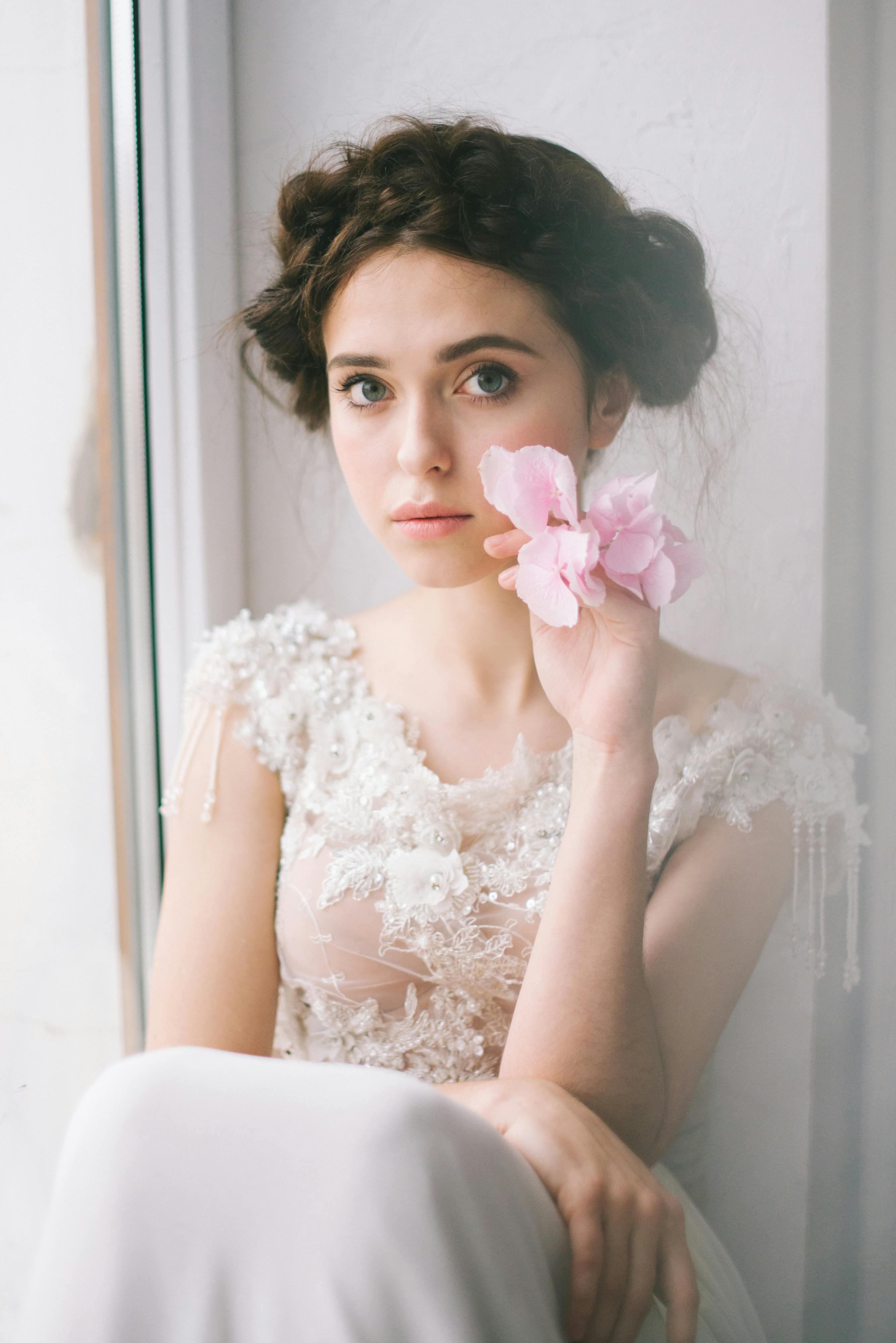 一位美丽的新娘穿着精致的连衣裙，在窗边摆了一朵粉红色的花