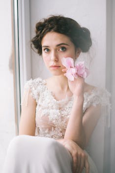 一位美丽的新娘穿着精致的连衣裙，在窗边摆了一朵粉红色的花