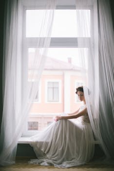 优雅的新娘穿着飘逸的礼服，坐在挂着透明窗帘的窗前，散发着优雅的气质