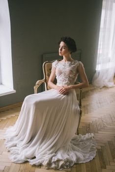 A serene bride in an elegant wedding dress poses indoors by a window.