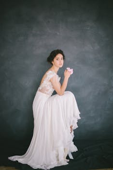 在黑暗的工作室背景下，优雅的新娘身着蕾丝礼服，手持淡粉色花朵摆着姿势