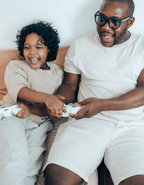 Free From above of positive African American father and child in light pajamas using gamepads while playing on game console together at home Stock Photo