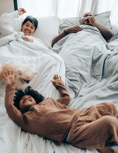 Free From above of black child in pajama playing with little dog while lying on bed of awake parents Stock Photo