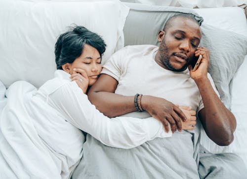From above of young African American man with closed eyes talking on smartphone lying in bed together with sleeping ethnic wife