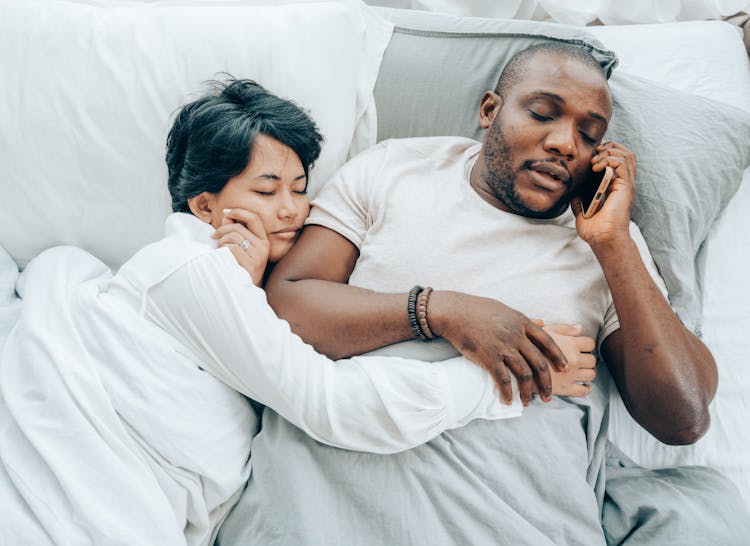 Asian Woman Sleeping Near Black Husband Talking On Smartphone