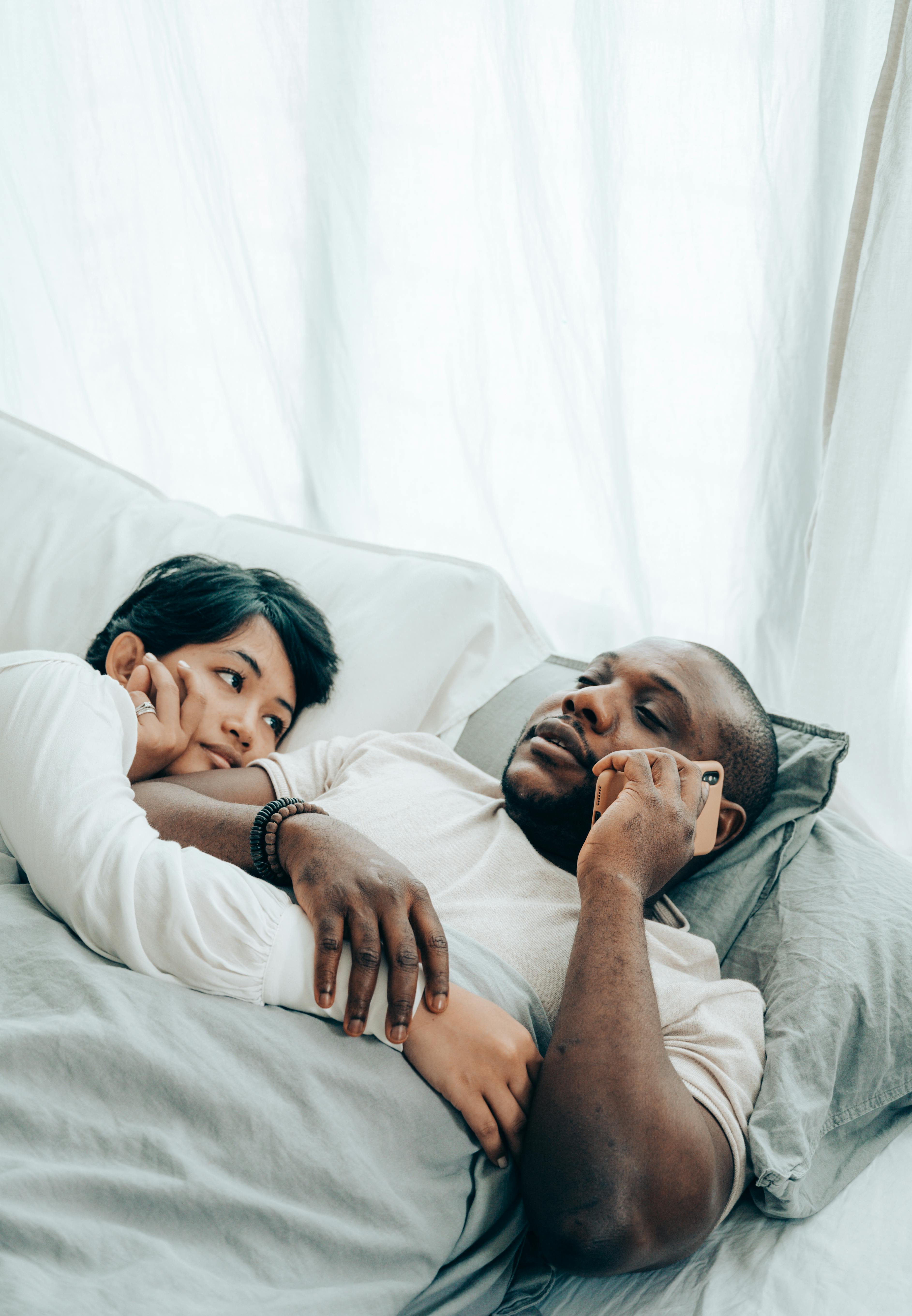 Young black man lying in bed with wife and talking on smartphone · Free Stock Photo