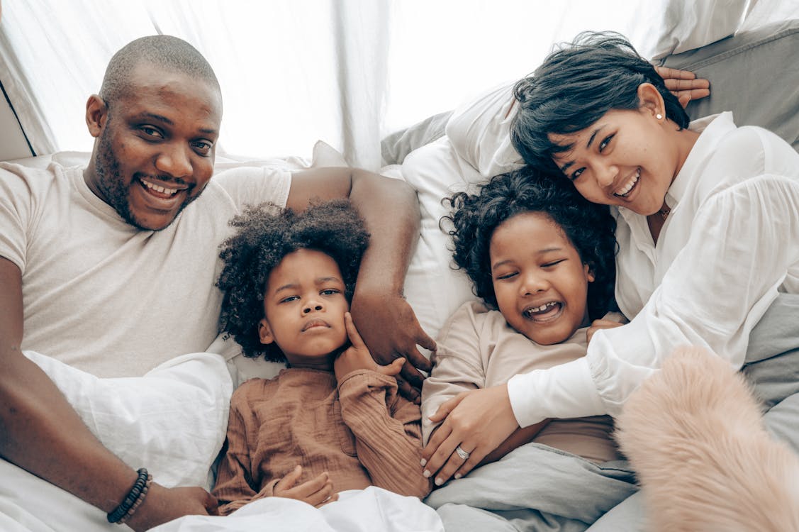 Cheerful multiracial parents with adorable little daughters hugging and smiling while sitting on comfortable bed in morning