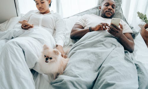 Crop pensive young couple lying in bed and using smartphone