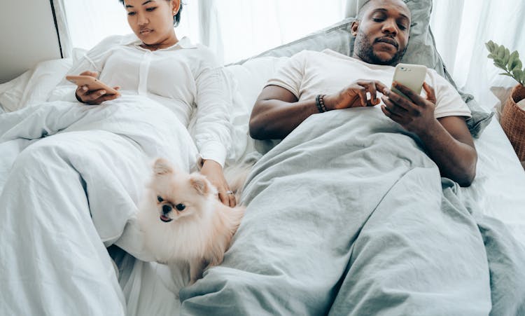 Crop Pensive Young Couple Lying In Bed And Using Smartphone