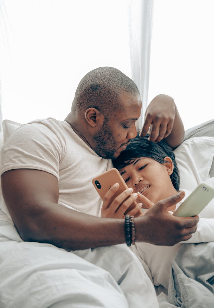 Loving Diverse Couple With Smartphones In Comfortable Bed