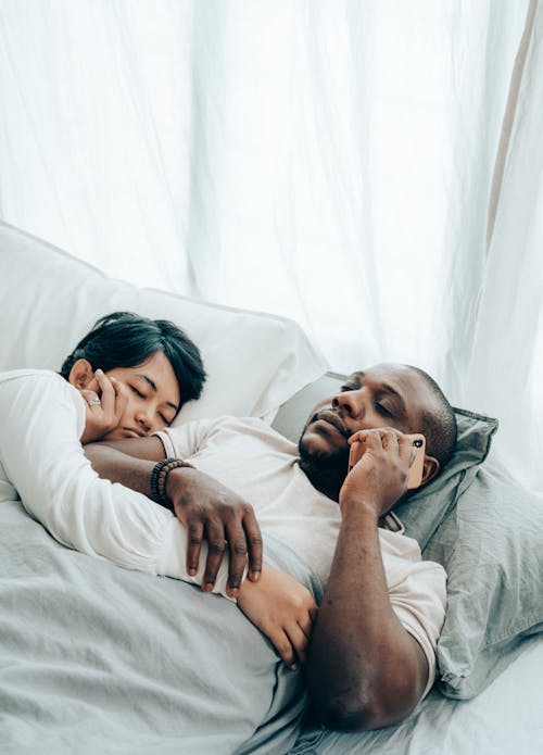 High angle of sleepy young black man lying in bed with closed eyes near wife and answering on phone call