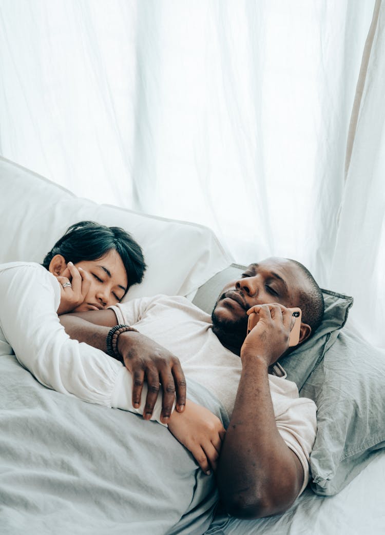 Sleeping Ethnic Woman Embracing Black Husband Lying And Talking On Smartphone