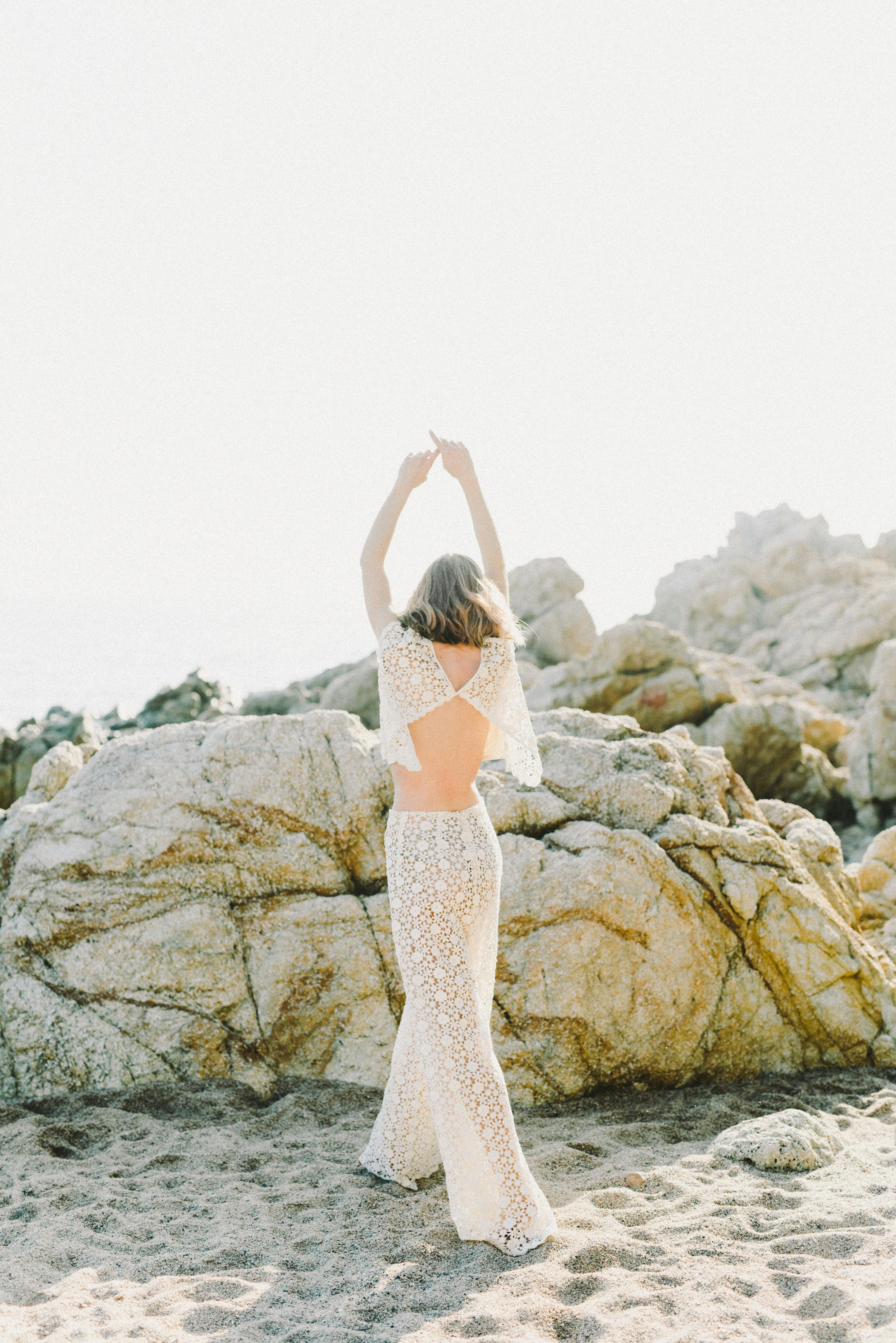 一名妇女穿着无背蕾丝连衣裙，双臂举起，站在阳光明媚的岩石海滩上