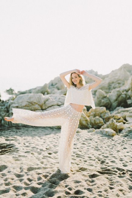 Elegant woman in a lace outfit poses gracefully on a sandy beach with rocky background.