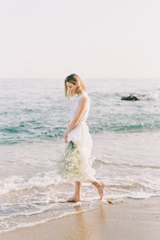 Woman in white dress walks barefoot along the beach, holding flowers.