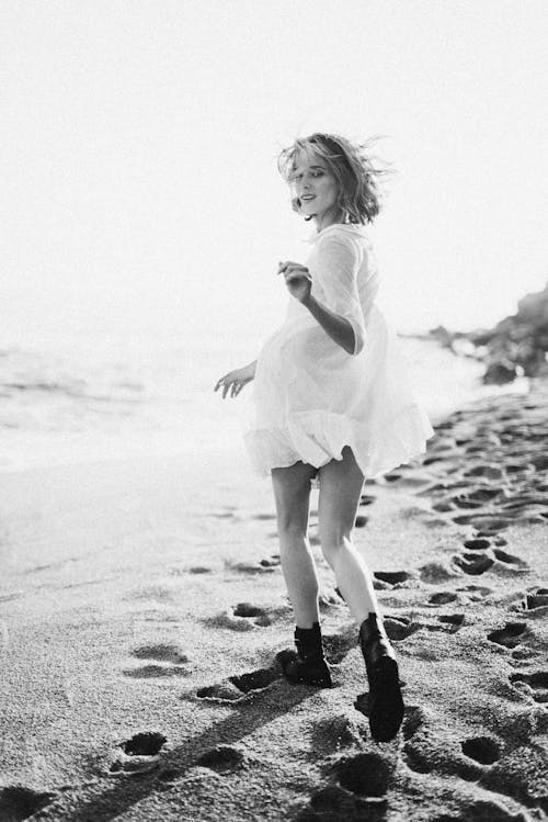 Grayscale Photo of Woman in White Dress Walking on Beach