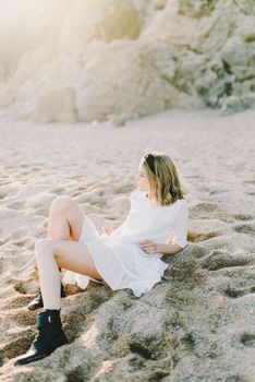 A woman in a white dress sits on a sandy beach by rock formations under the sun.