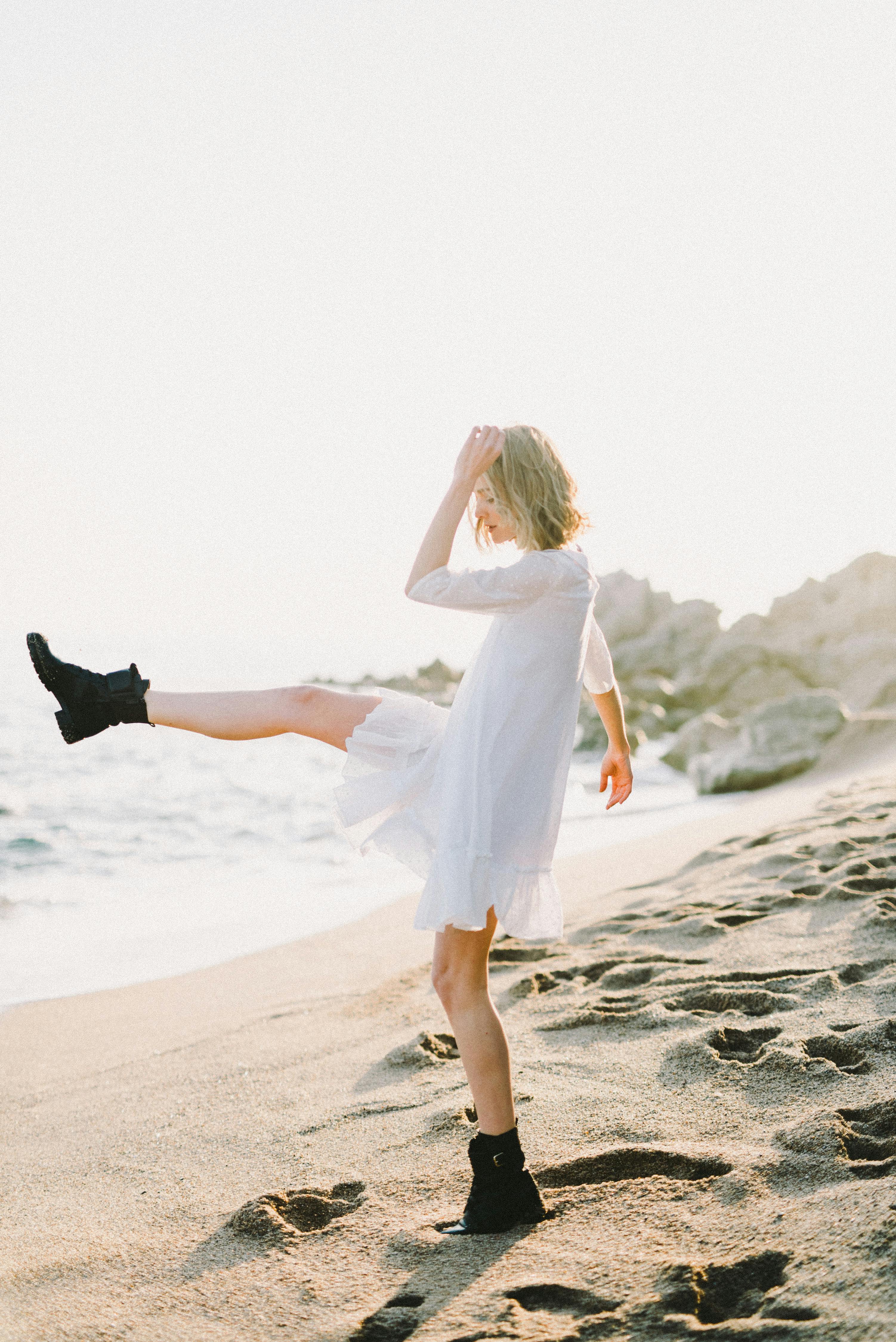 一个穿着白色连衣裙和黑色靴子的女人，在阳光明媚的海滩上嬉戏地踢着沙子