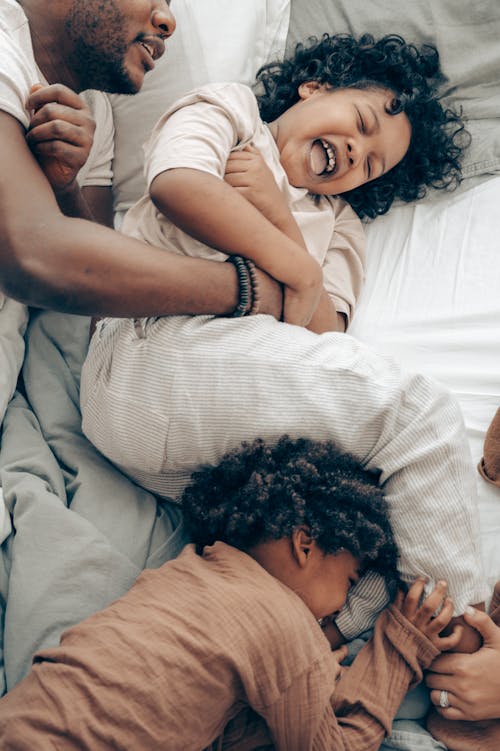 From above of crop black father tickling and playing with adorable laughing daughters while lying on bed in morning