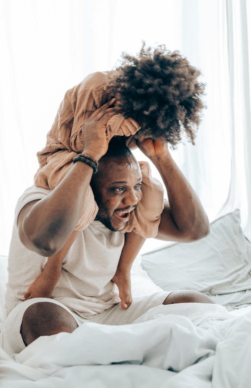 Cheerful African American man with kid in casual clothes hugging and playing while sitting on bed in comfortable bedroom on weekend
