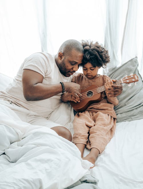 Free From above of focused African American guy and child with curly hair sitting on bed and playing on guitar during weekend Stock Photo