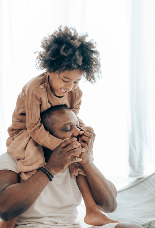 Free African American father kissing hands of kid in casual wear while having fun together in cozy living room during weekend day Stock Photo