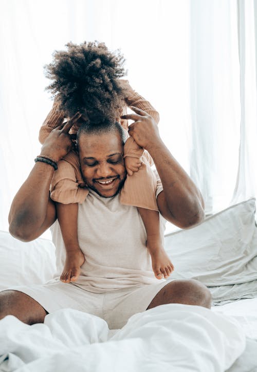 African American kid with curly hair sitting around neck of parent and kissing in head while sitting on bed and having fun together on daytime