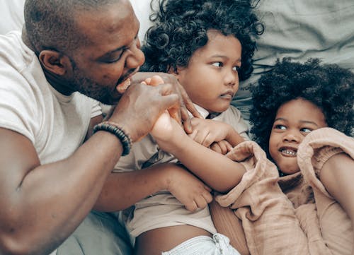 Free From above of African American man embracing with adorable children on cozy bed while chilling and having fun together at home Stock Photo