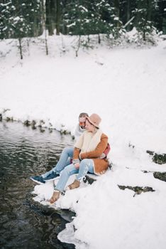在白雪皑皑的冬季公园里，一对夫妇坐在河边相拥