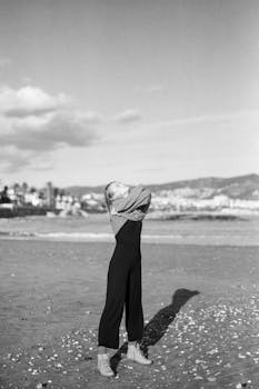 A woman stretches on a beach, wearing a sweater, in a black and white photo.
