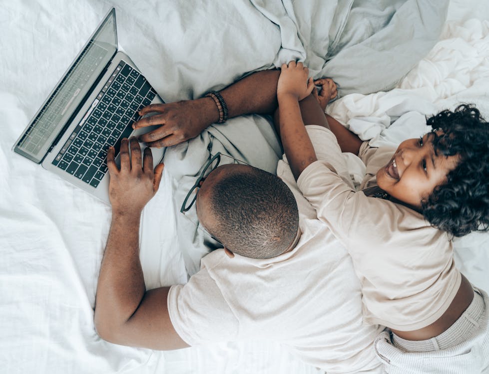 Free Top view of cheerful African American kid lying near father while man typing on keyboard of laptop during remote work in cozy bedroom Stock Photo