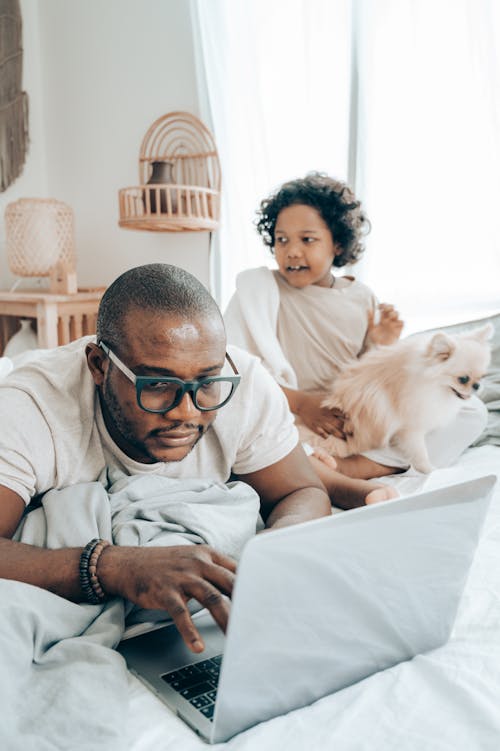 African American child with white small dog sitting behind father while man browsing laptop on bed in cozy apartment on daytime