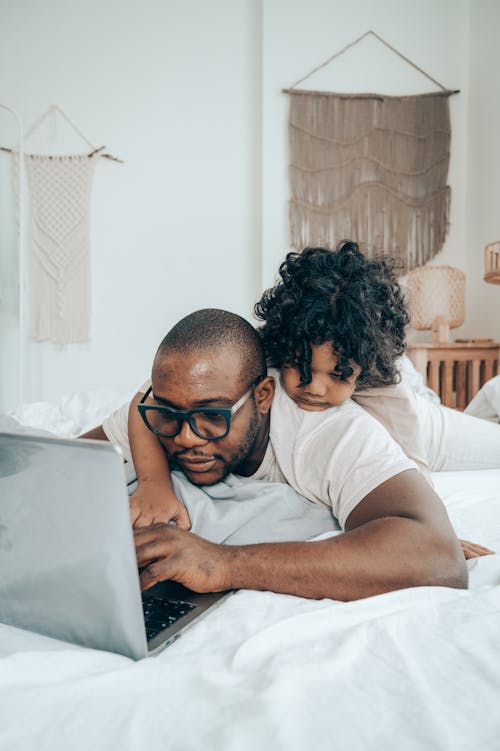Serious African American kid with curly hair lying on back of father while browsing laptop together during weekend on daytime