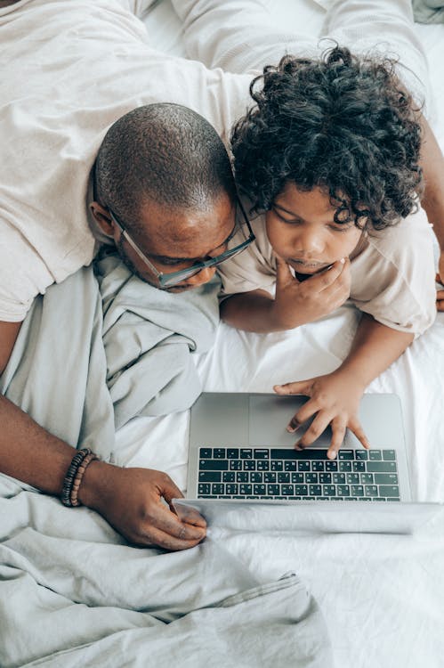 From above of thoughtful black male in eyeglasses with child with curly hair lying on bed and using laptop