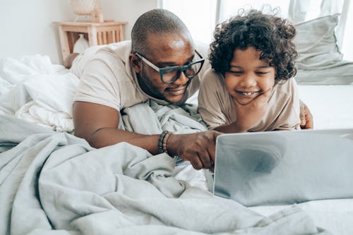 Cheerful African American male in glasses lying on bed in bedroom with child in sleepwear and smiling while browsing laptop