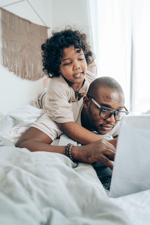 African American parent in glasses and kid with curly hair in pajama lying on bed in bedroom and browsing laptop