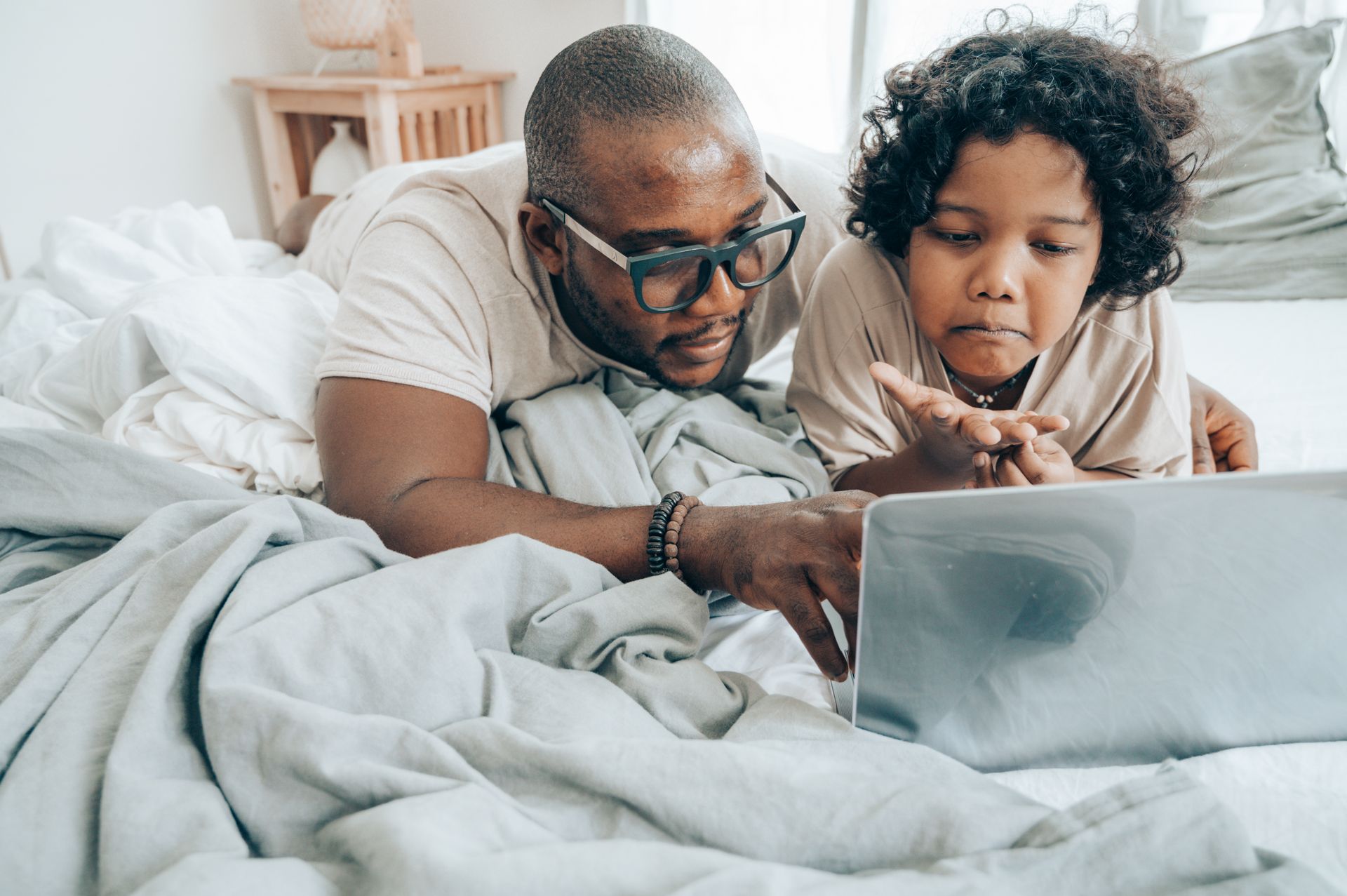 A father and child lying on bed, using a laptop for online activities, in a cozy home setting.