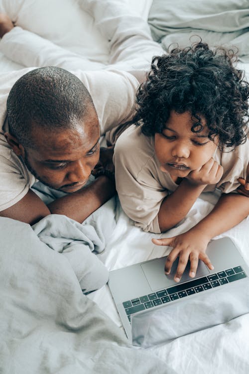 From above of concentrated African American kid with curly hair and father lying on bed in bedroom and browsing laptop