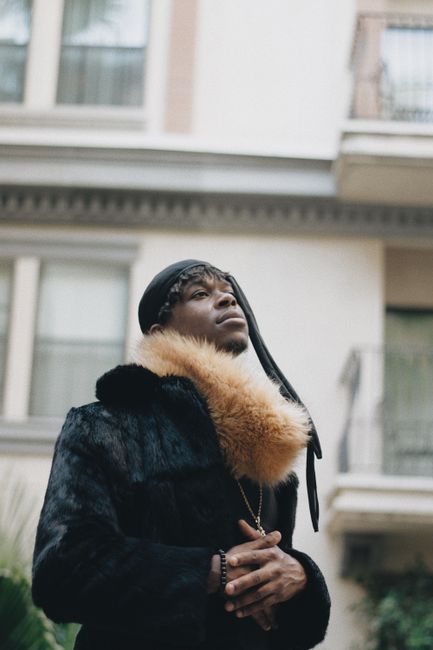 A stylish man in a fur coat poses outdoors against an urban Los Angeles backdrop.